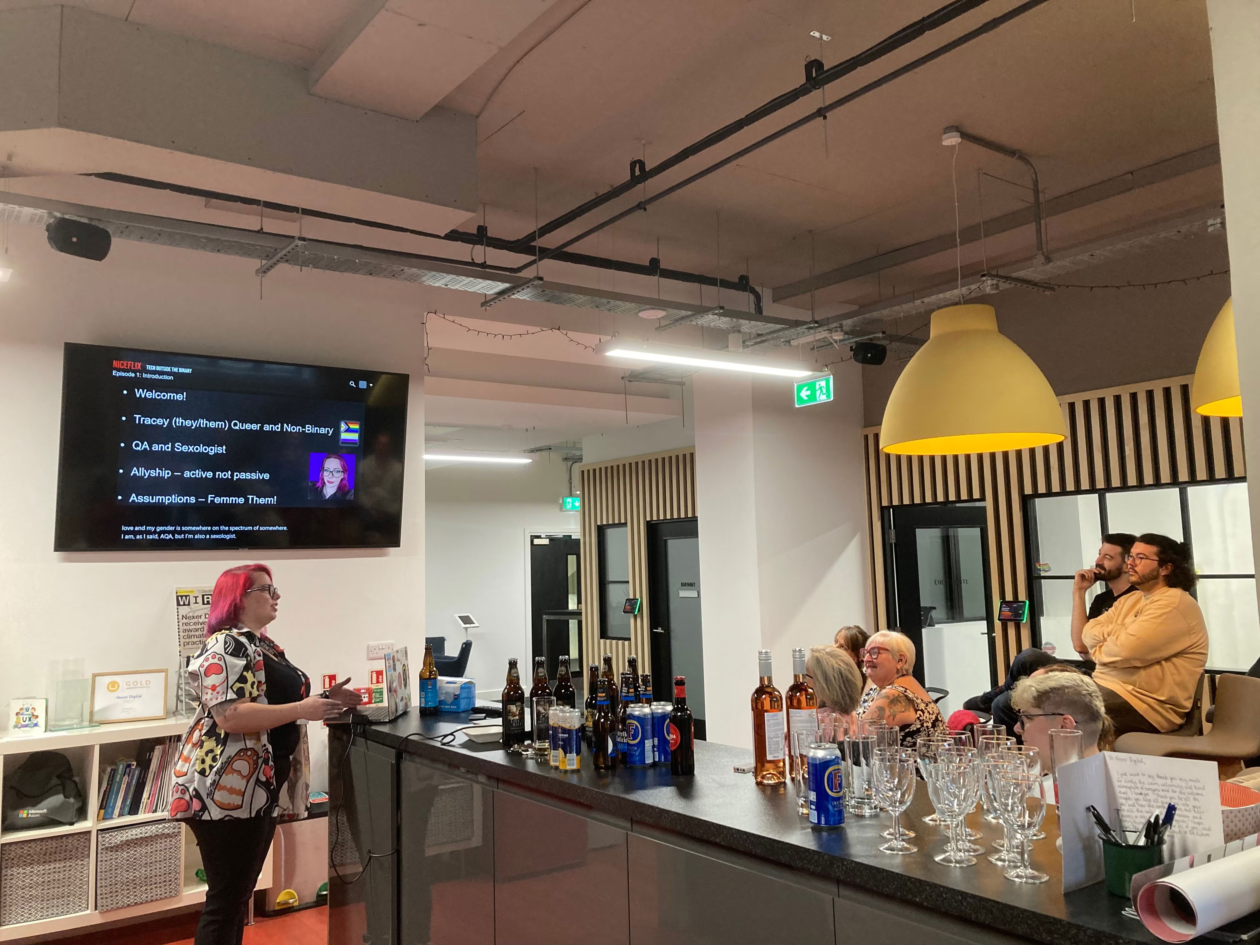 Tracey presenting to an audience. Presentation on screen to the side, audience to the front. In between is a table of drinks, and a the computer in use